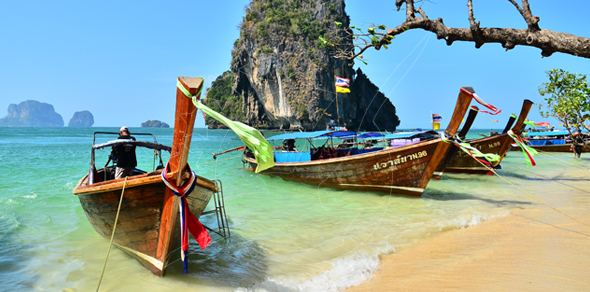 Bateaux à longue queue en bois amarrés sur une plage de sable thaïlandaise, eau turquoise et falaise karstique en fond