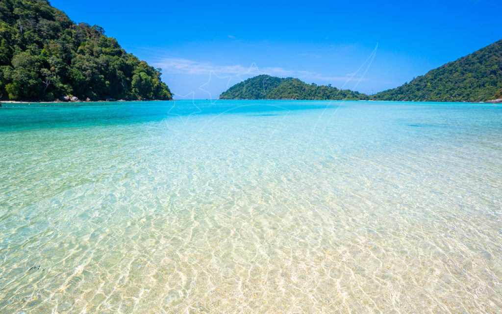 Aguas turquesas cristalinas sobre arena clara con islas cubiertas de jungla y cielo azul, bahía tropical