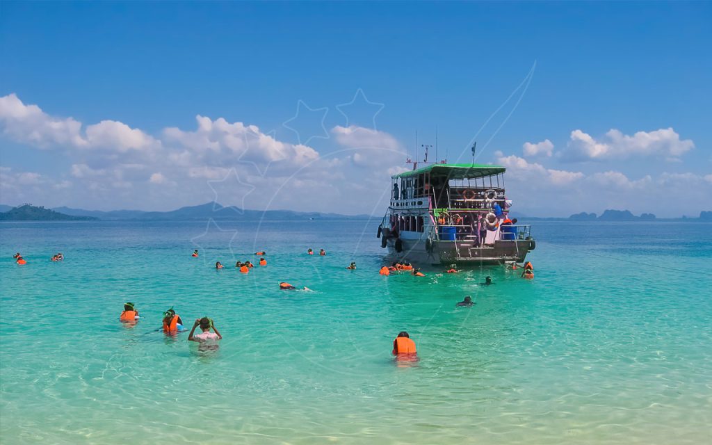นักดำน้ำตื้นสวมเสื้อชูชีพสีส้มลอยตัวในน้ำตื้นใสสีเทอร์คอยส์ข้างเรือทัวร์ไม้นอกเกาะเขตร้อน