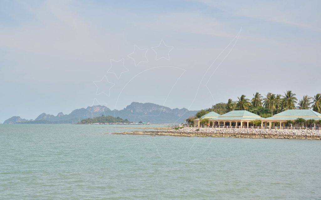 Pavilhões de telhado verde e palmeiras num quebra-mar rochoso, com colinas calcárias do outro lado do mar calmo e enevoado