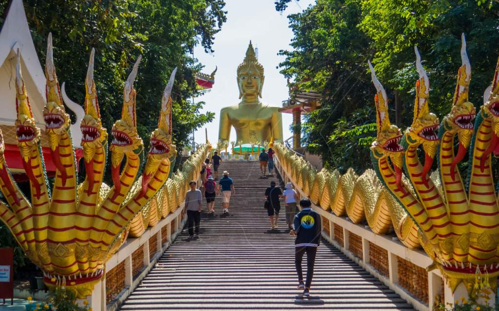 Goldene Naga-Schlangen als Geländer an einer Tempeltreppe hinauf zu einer großen sitzenden goldenen Buddha-Statue, Thailand