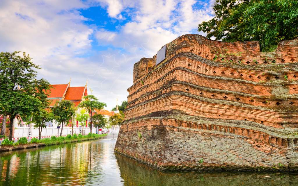 Angolo di mattoni rossi delle antiche mura di Chiang Mai accanto al fossato verde, con tetto di un tempio e alberi