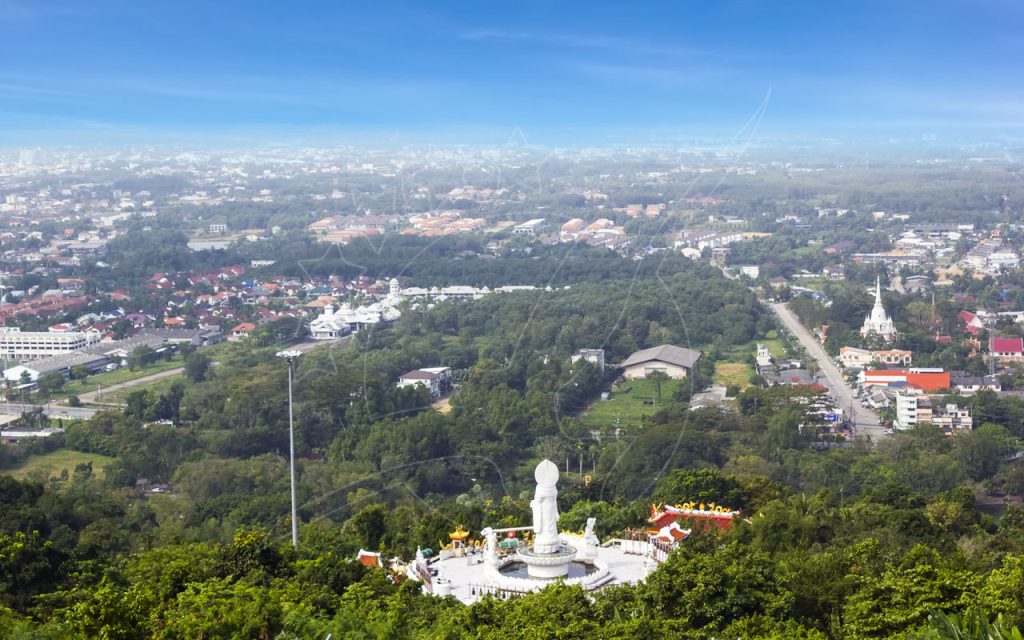 Weiße Guanyin-Statue und Schrein auf Hügelkuppe über bewaldeten Hängen mit Blick auf thailändische Stadt und Tempeltürme