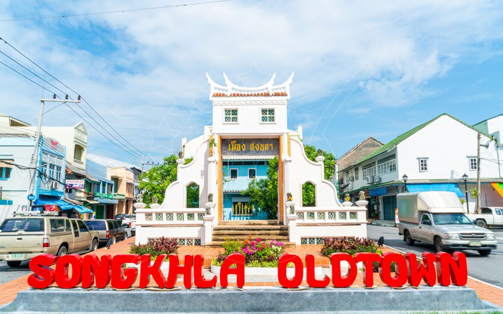 Rote Buchstaben „SONGKHLA OLDTOWN“ vor einem weißen chinesischen Torbogen in einer sonnigen thailändischen Straße