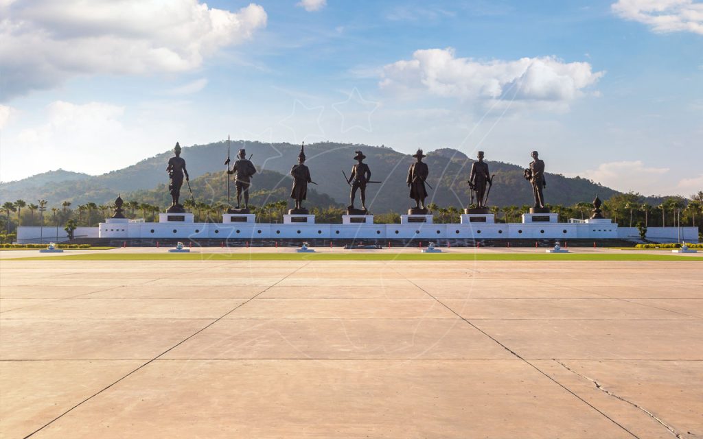 Siete estatuas de bronce de reyes sobre un muro-pedestal blanco en el parque Rajabhakti, Hua Hin, con colinas verdes detrás