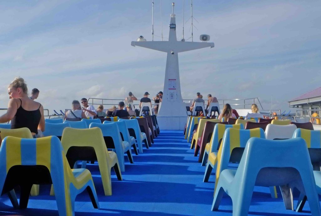 Offenes Fährdeck mit blau-gelben Sitzen, Passagieren und Stahlkonstruktion vor bewölktem Himmel